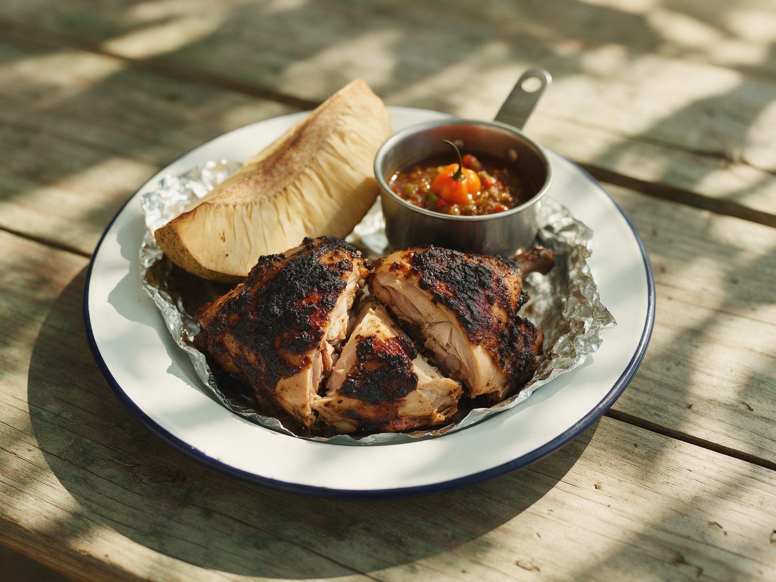 A chopped jerk chicken quarter with charred skin on foil, with roast breadfruit and a small pot of scotch bonnet sauce.